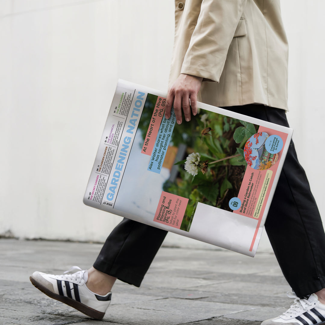 A person in a beige trench coat, black trousers, and white Adidas sneakers walks while carrying a large printed gardening magazine or poster titled 'GARDENING NATION' with colorful text and a flower photo.